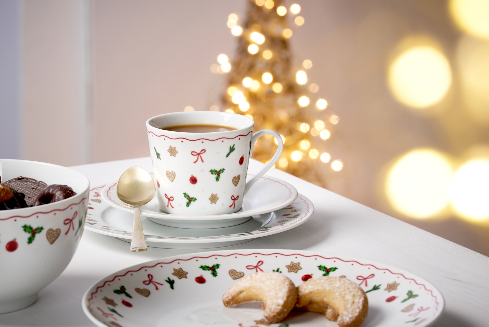Kerstversierd koffieservies met kopje, schoteltje en bord, op de voorgrond een gouden lepel, op de achtergrond een wazige kerstboom, zijaanzicht.