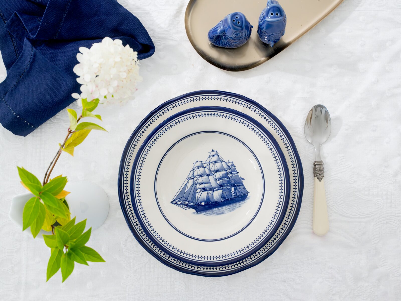 Draufsicht auf blau-weißes Porzellan-Tellerset mit Segelschiff-Motiv und dekorativem Rand, auf weißer Tischdecke arrangiert mit Löffel, blauer Serviette, Blume und Salz-und-Pfefferstreuern.