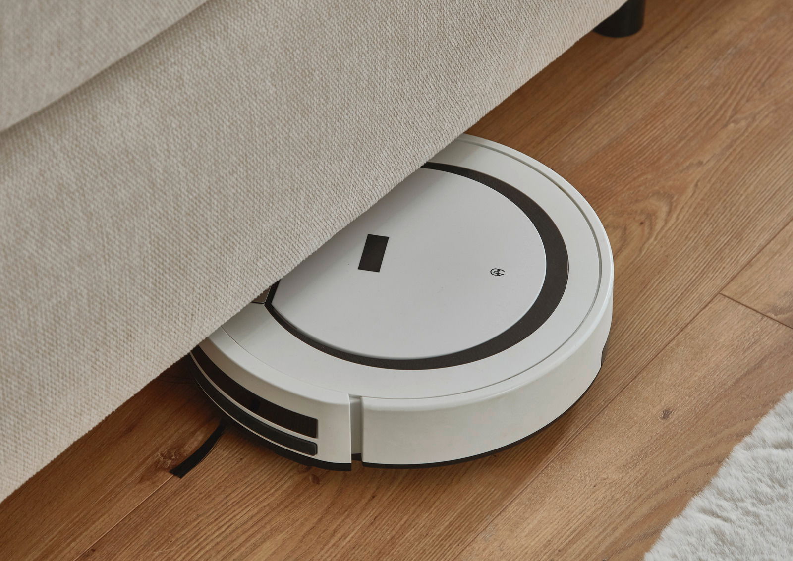 Ein weißer Saugroboter reinigt den Boden unter einem Boxspringbett aus der Seitenperspektive.