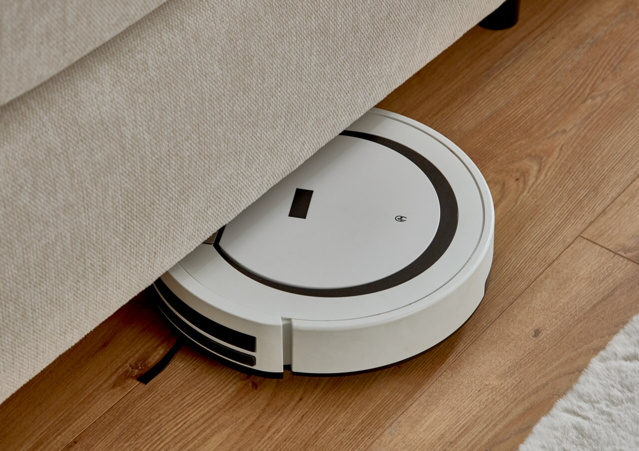 Ein weißer Saugroboter reinigt den Boden unter einem Boxspringbett aus der Seitenperspektive.