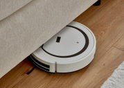 Een witte robotstofzuiger maakt de vloer onder een boxspringbed schoon, gezien vanuit de zijkant.