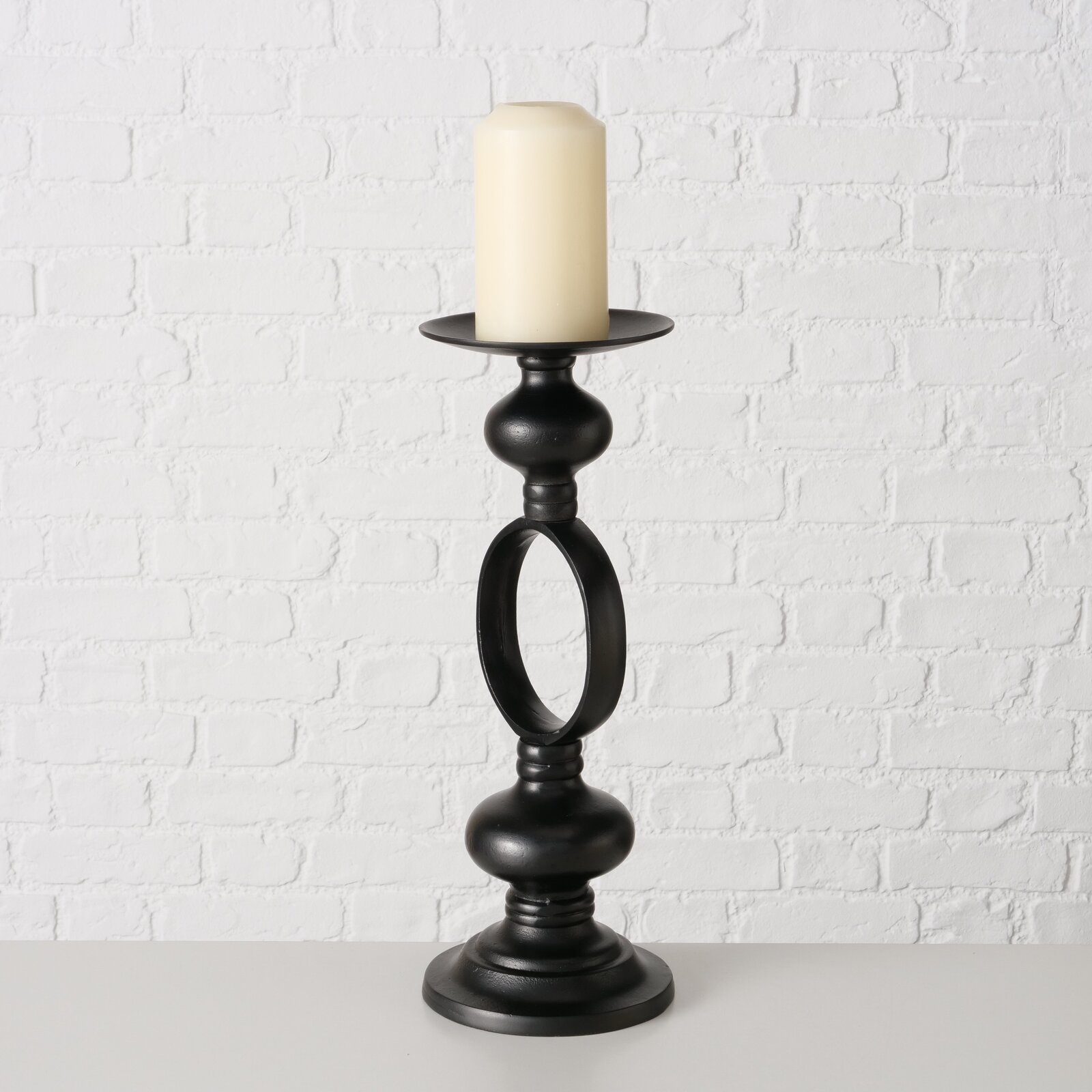 Schwarzer Kerzenleuchter Plokky mit einer cremefarbenen Kerze, frontal vor einer weißen Ziegelwand fotografiert.