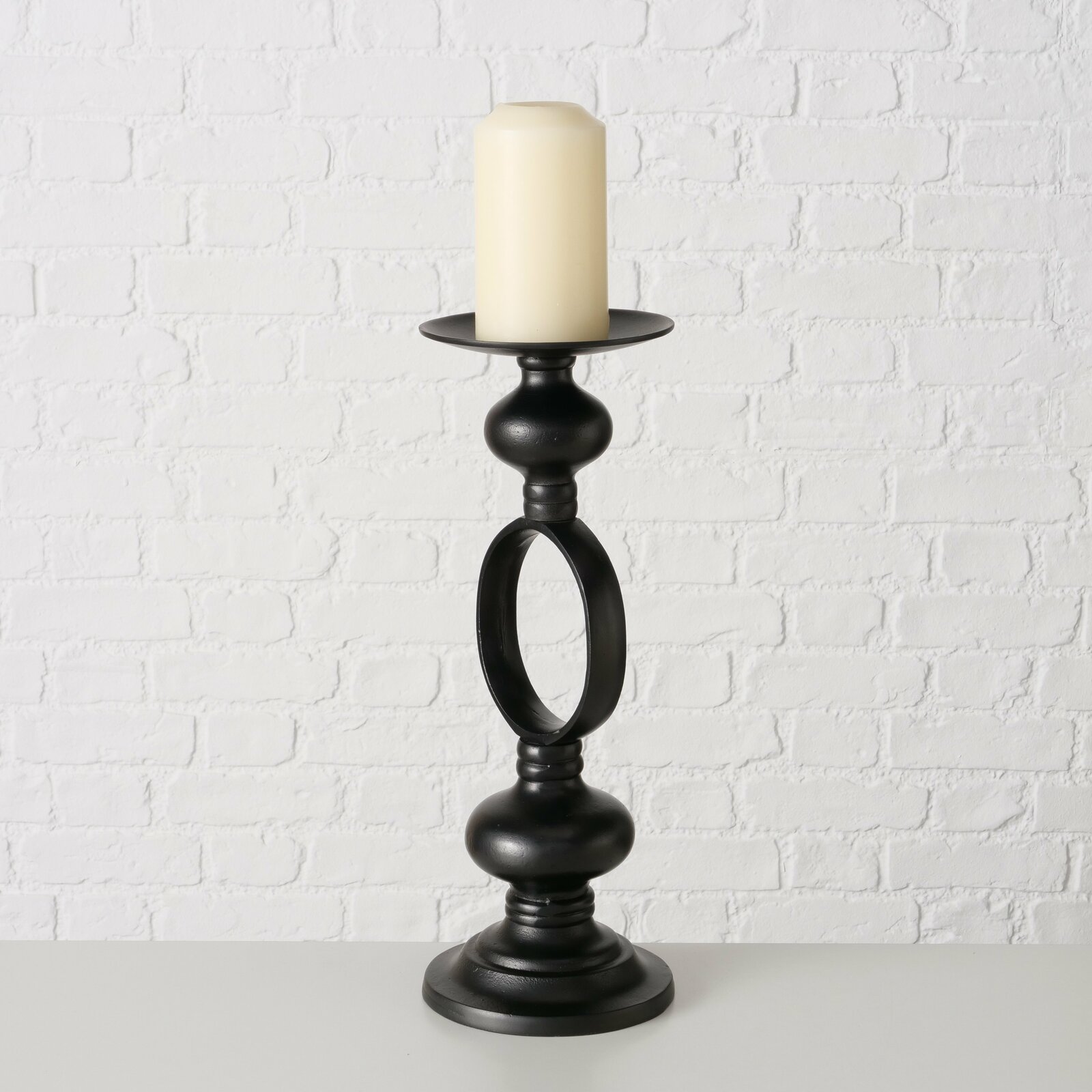 Schwarzer Kerzenleuchter Plokky mit einer cremefarbenen Kerze, frontal vor einer weißen Ziegelwand fotografiert.