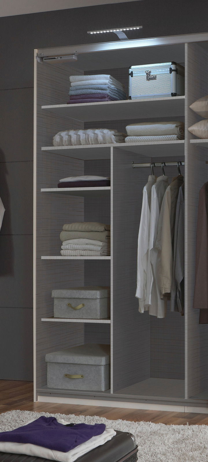 Ein geöffneter Kleiderschrank mit mehreren Regalböden, auf denen gefaltete Kleidung und Boxen liegen, sowie einer Kleiderstange mit hängenden Hemden. Die Perspektive zeigt den Schrank von vorne.