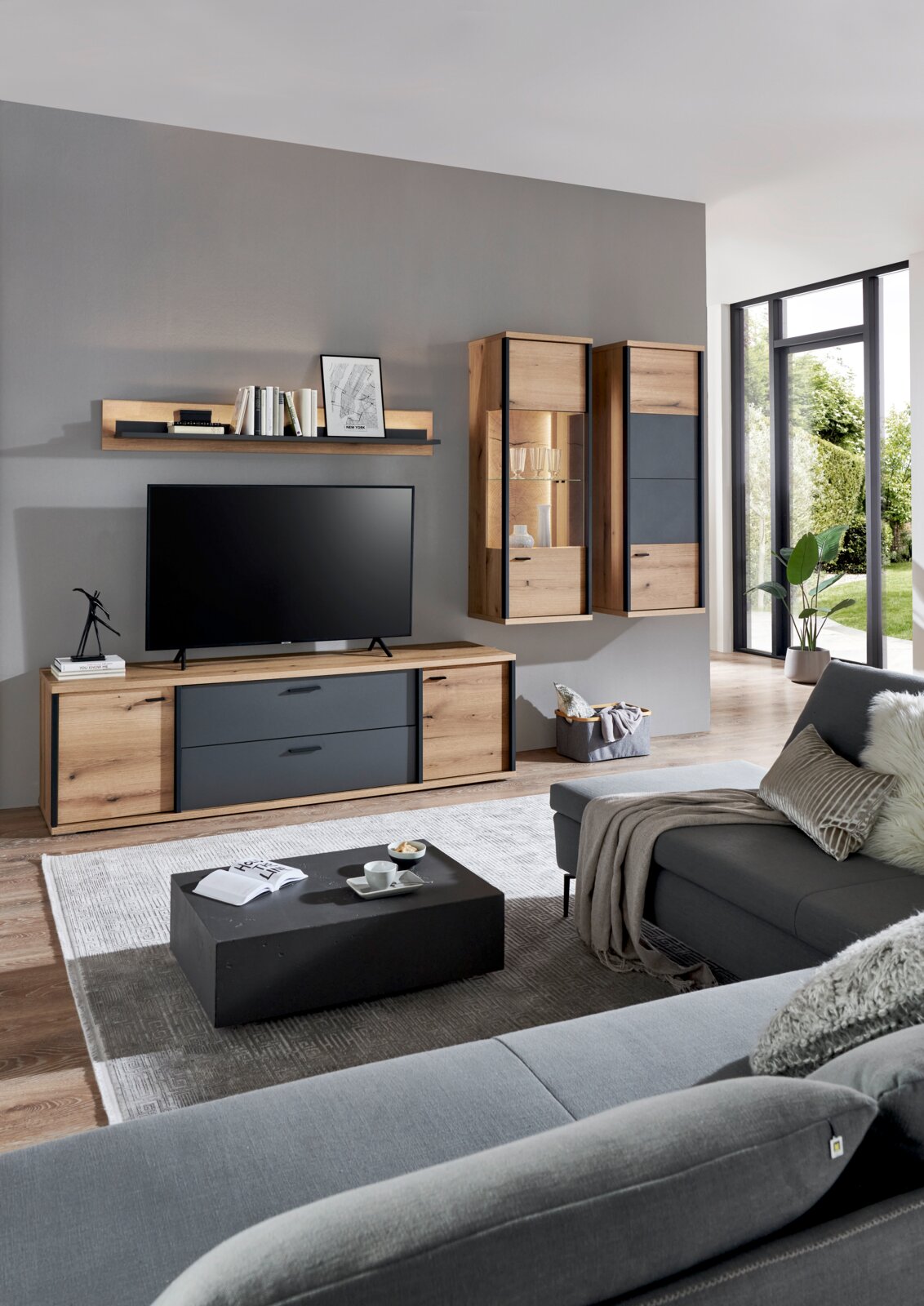 Modernes Wohnzimmer mit TV-Lowboard in Holz- und Anthrazitoptik, zwei hängenden Vitrinen und Wandregal an grauer Wand; Perspektive: frontale Ansicht mit leichtem Blick von rechts