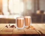 Zwei Thermogläser mit Cappuccino auf einem Holztisch, umgeben von Kaffeebohnen und Gebäck, aus frontaler Perspektive fotografiert.