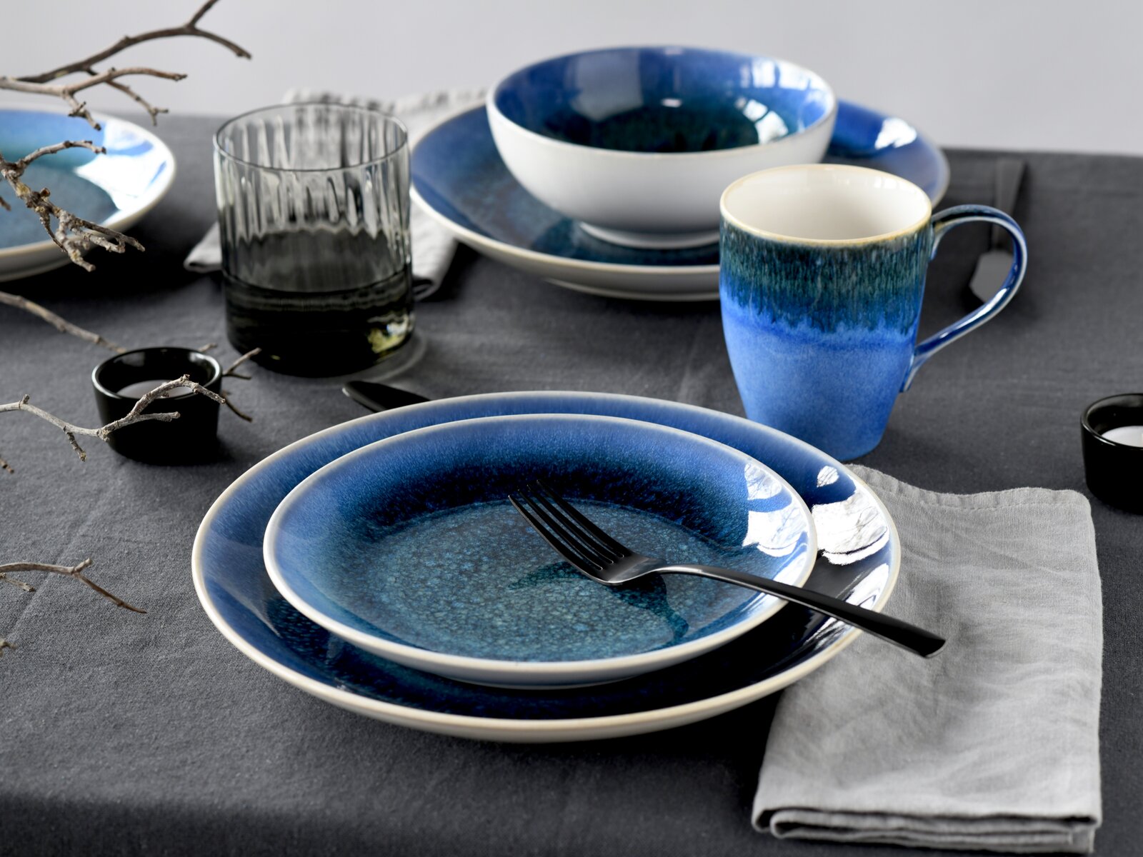 Gedeckter Tisch mit blau glasiertem Keramikgeschirr (Teller, Schüssel und Tasse) sowie Glas und Besteck auf grauem Tuch, fotografiert aus leicht erhöhter schräger Perspektive (3/4-Ansicht).