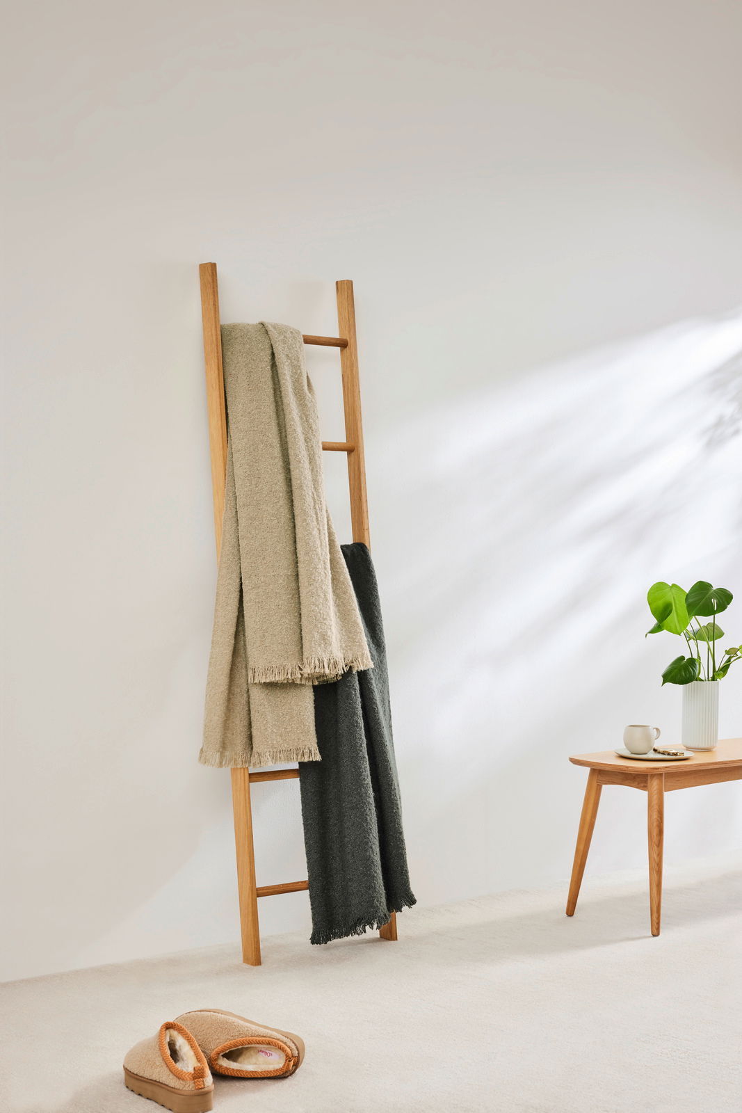 Zwei Wohndecken in Beige und Dunkelgrau hängen auf einer Holzleiter in einem hellen Raum. Perspektive von vorne.