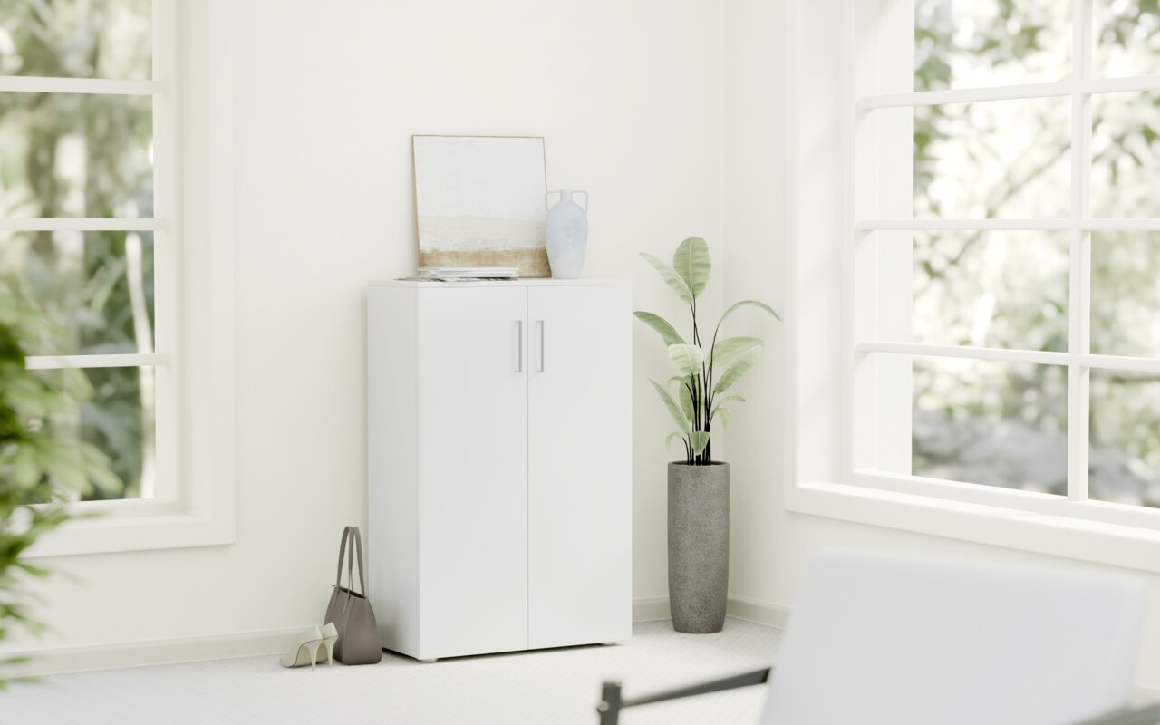 Weißer, niedriger Schuhschrank mit zwei Türen, dekoriert mit einer Vase und einem Bild, in einem hellen Raum mit großen Fenstern, seitliche Perspektive.