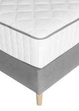Nahaufnahme eines Boxspringbetts mit weißer, gesteppten Matratze und grauem Unterbau auf Holzfüßen, Perspektive von der Ecke.
