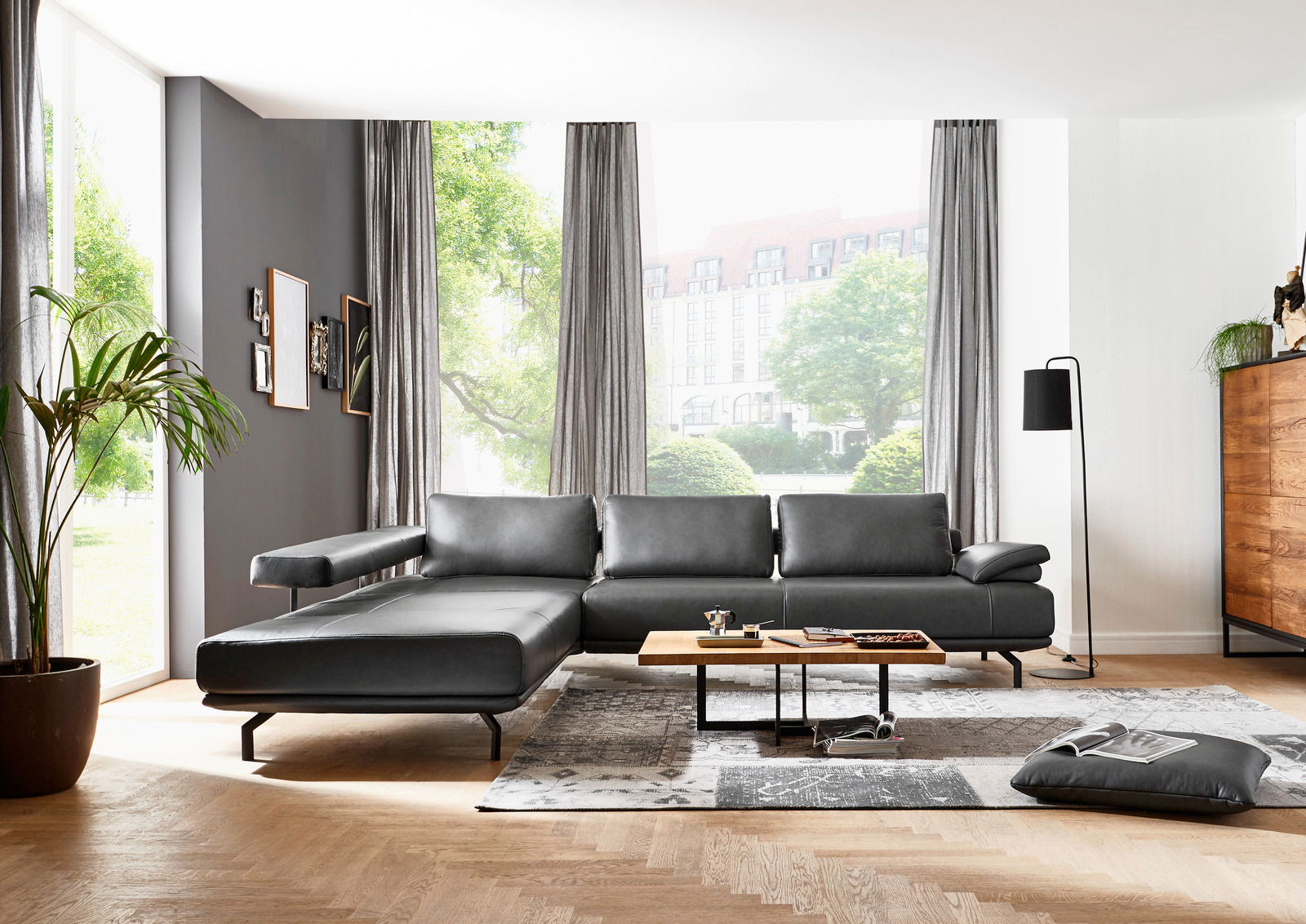 Moderne graue Ledercouch in einem stilvollen Wohnzimmer mit großen Fenstern, die viel Tageslicht hereinlassen. Perspektive von vorne links.