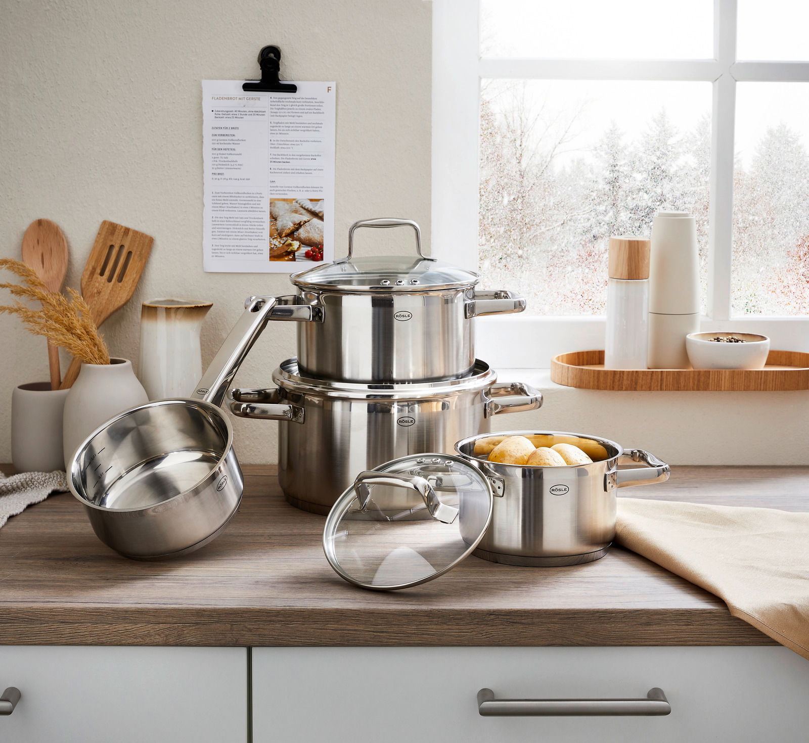 Topfset Charm 4-tlg. aus Edelstahl, bestehend aus drei Töpfen und einer Stielkasserolle, auf einer Küchenarbeitsplatte vor einem Fenster mit winterlicher Aussicht. Perspektive: Frontalansicht.