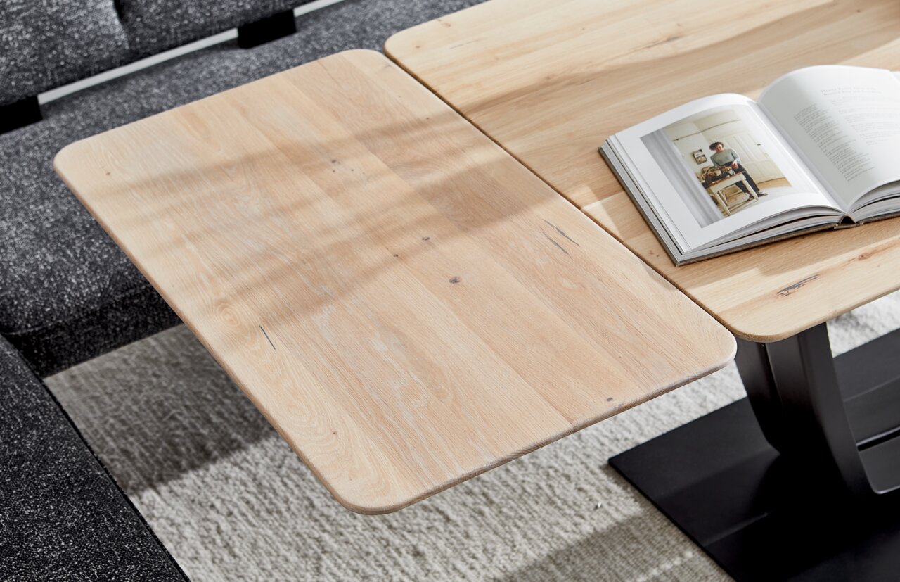 Nahaufnahme eines Esstisches aus hellem Holz mit abgerundeten Ecken, auf dem ein aufgeschlagenes Buch liegt. Der Tisch steht auf einem dunklen Teppich und ist von einer grauen Couch umgeben.
