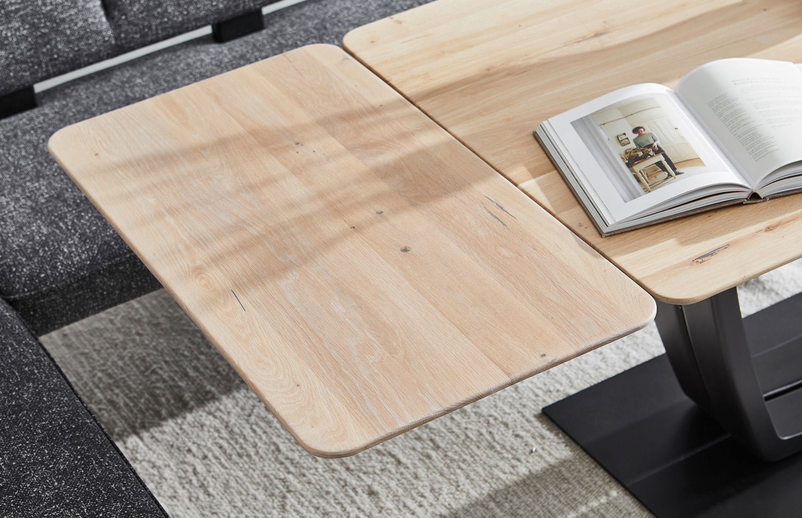 Nahaufnahme eines Esstisches aus hellem Holz mit abgerundeten Ecken, auf dem ein aufgeschlagenes Buch liegt. Der Tisch steht auf einem dunklen Teppich und ist von einer grauen Couch umgeben.