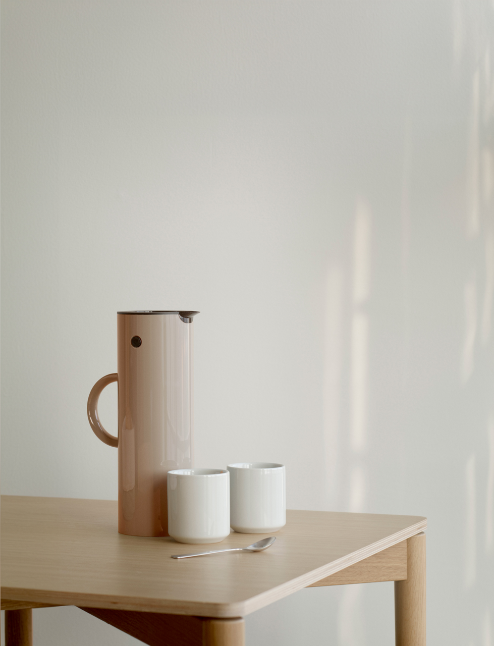 Stelton Thermoskan Seitliche Ansicht einer EM77 Isolierkanne in Weizenfarbe auf einem Holztisch, begleitet von zwei weißen Tassen und einem Löffel.