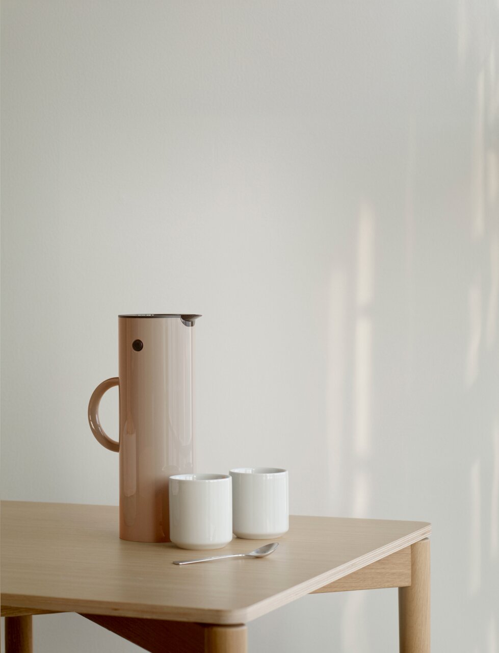 Stelton Isolierkanne Seitliche Ansicht einer EM77 Isolierkanne in Weizenfarbe auf einem Holztisch, begleitet von zwei weißen Tassen und einem Löffel.