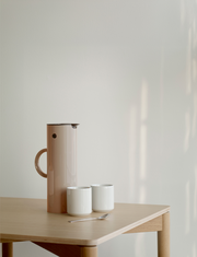 Seitliche Ansicht einer EM77 Isolierkanne in Weizenfarbe auf einem Holztisch, begleitet von zwei weißen Tassen und einem Löffel.