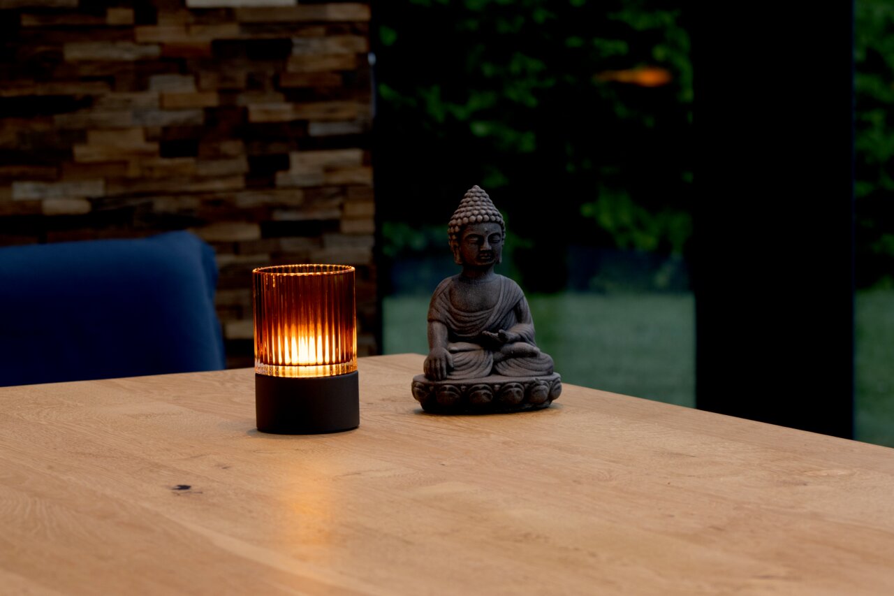 Fischer & Honsel Akku-Windlicht ARIVA PRO Akku-Windlicht auf einem Holztisch neben einer Buddha-Statue, mit warmem Licht in einem modernen Raum.