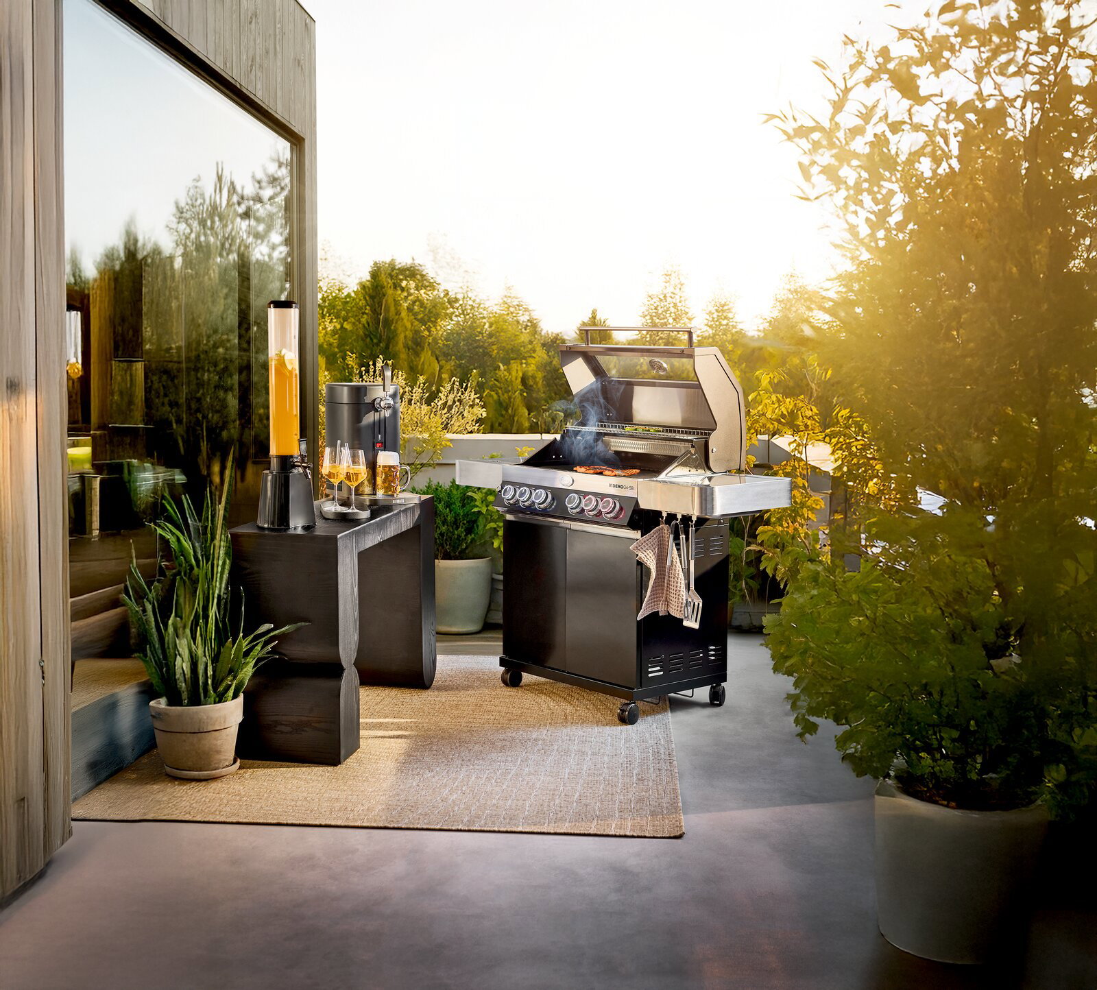 Edelstahl-Gasgrill mit geöffnetem Deckel und Seitenablagen auf moderner Terrasse im Abendlicht, frontale leicht seitliche Perspektive