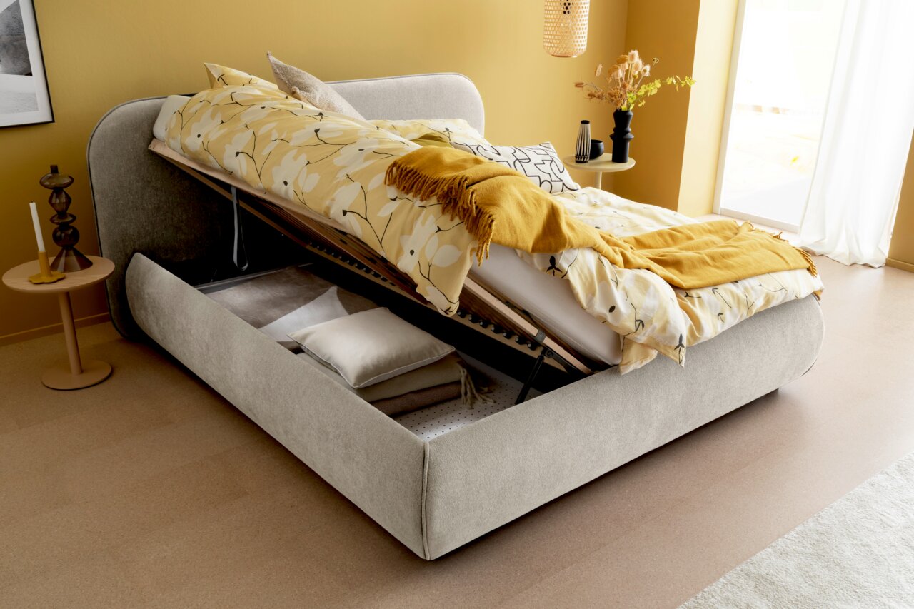 Polsterbett LISSABON mit hochgeklappter Matratze, zeigt Stauraum darunter, seitliche Perspektive, gelbe Bettwäsche und Kissen.