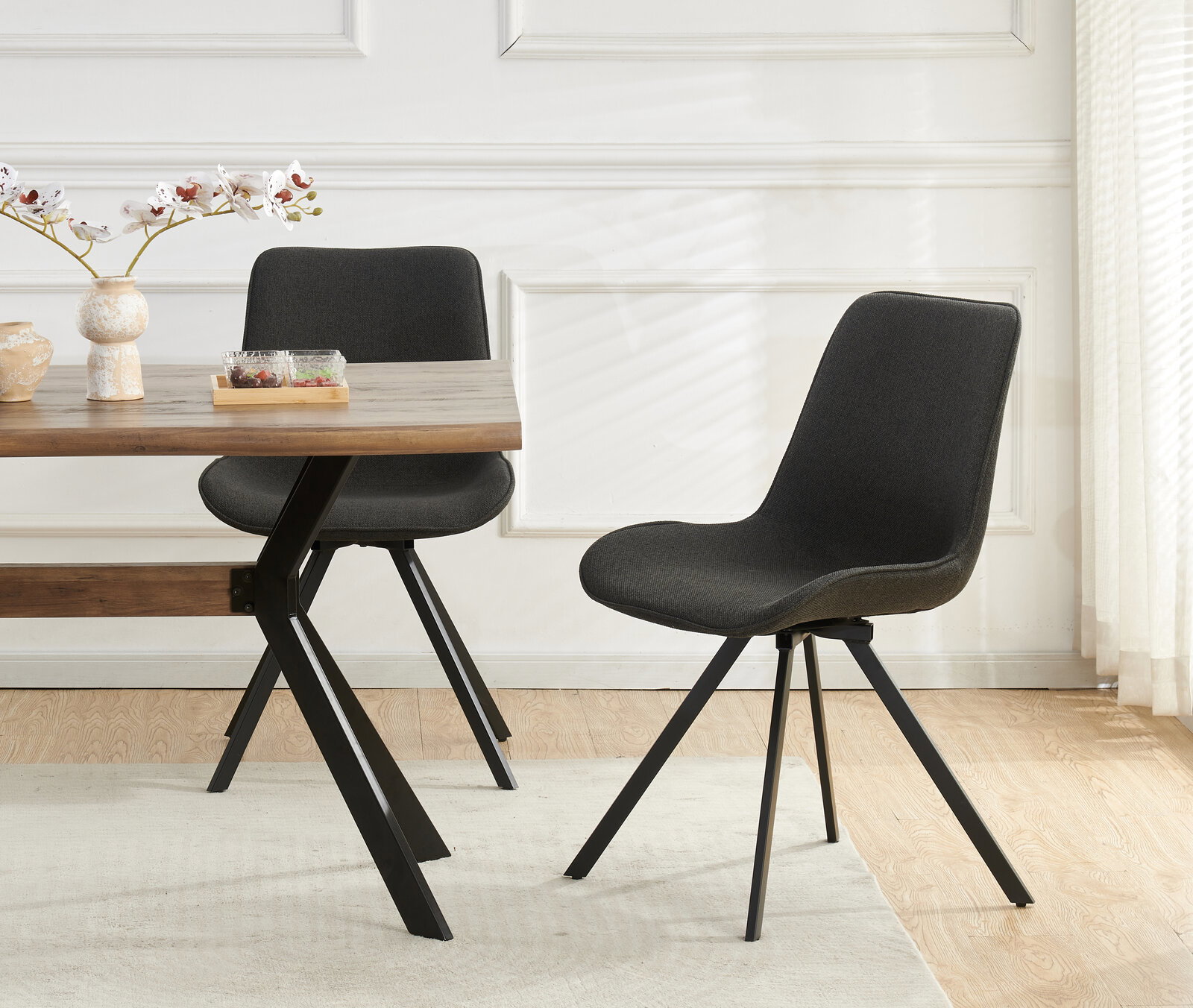Schwarzer, gepolsterter Esszimmerstuhl mit Stoffbezug und schwarzen Metallbeinen neben einem Holztisch in hellem Raum, seitliche Perspektive