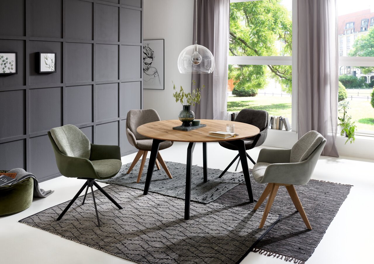 Runder Esstisch mit Holzplatte und schwarzen Metallbeinen, umgeben von vier gepolsterten Stühlen in einem modernen Esszimmer mit großen Fenstern und grauer Wand. Perspektive: seitlich.