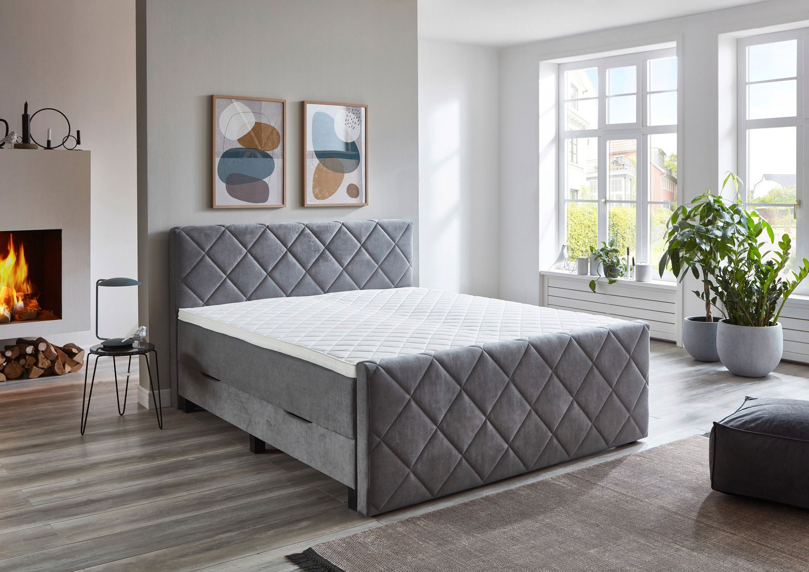 Graues Boxspringbett BX2200 CHESTER in einem modernen Schlafzimmer mit Kamin und großen Fenstern, seitliche Perspektive.