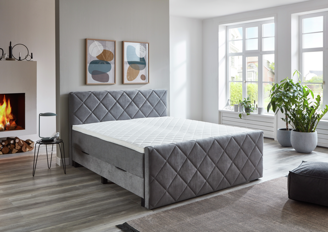 Graues Boxspringbett BX2200 CHESTER in einem modernen Schlafzimmer mit Kamin und großen Fenstern, seitliche Perspektive.