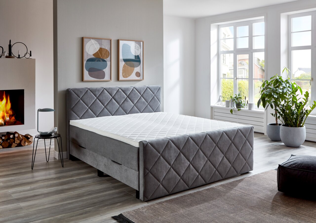 Graues Boxspringbett BX2200 CHESTER in einem modernen Schlafzimmer mit Kamin und großen Fenstern, seitliche Perspektive.