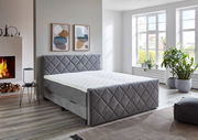 Graues Boxspringbett BX2200 CHESTER in einem modernen Schlafzimmer mit Kamin und großen Fenstern, seitliche Perspektive.
