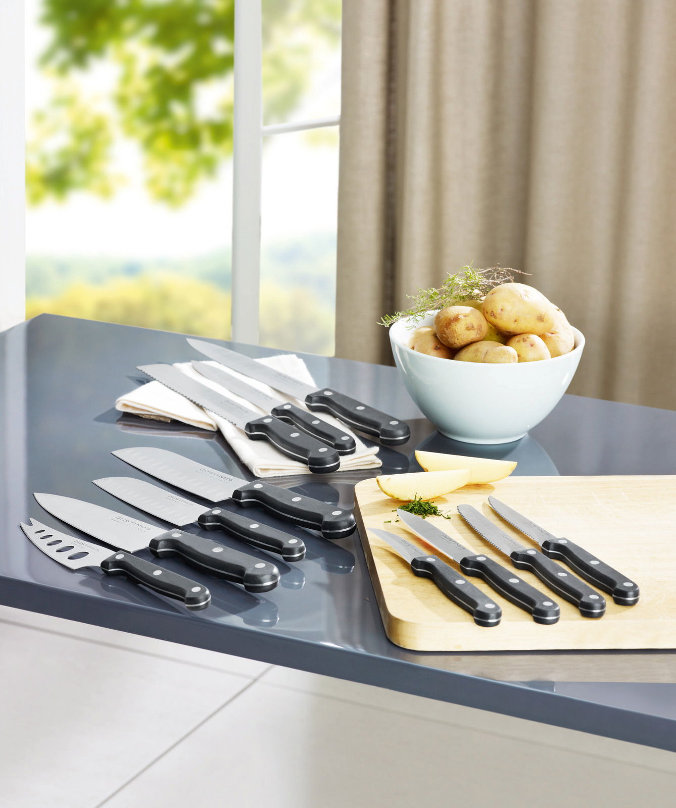 Justinus Brötchenmesser Küchenchef KÜCHENCHEF Verschiedene Küchenmesser, darunter ein Brötchenmesser, auf einem Tisch in einer modernen Küche, mit Blick auf ein Fenster im Hintergrund.