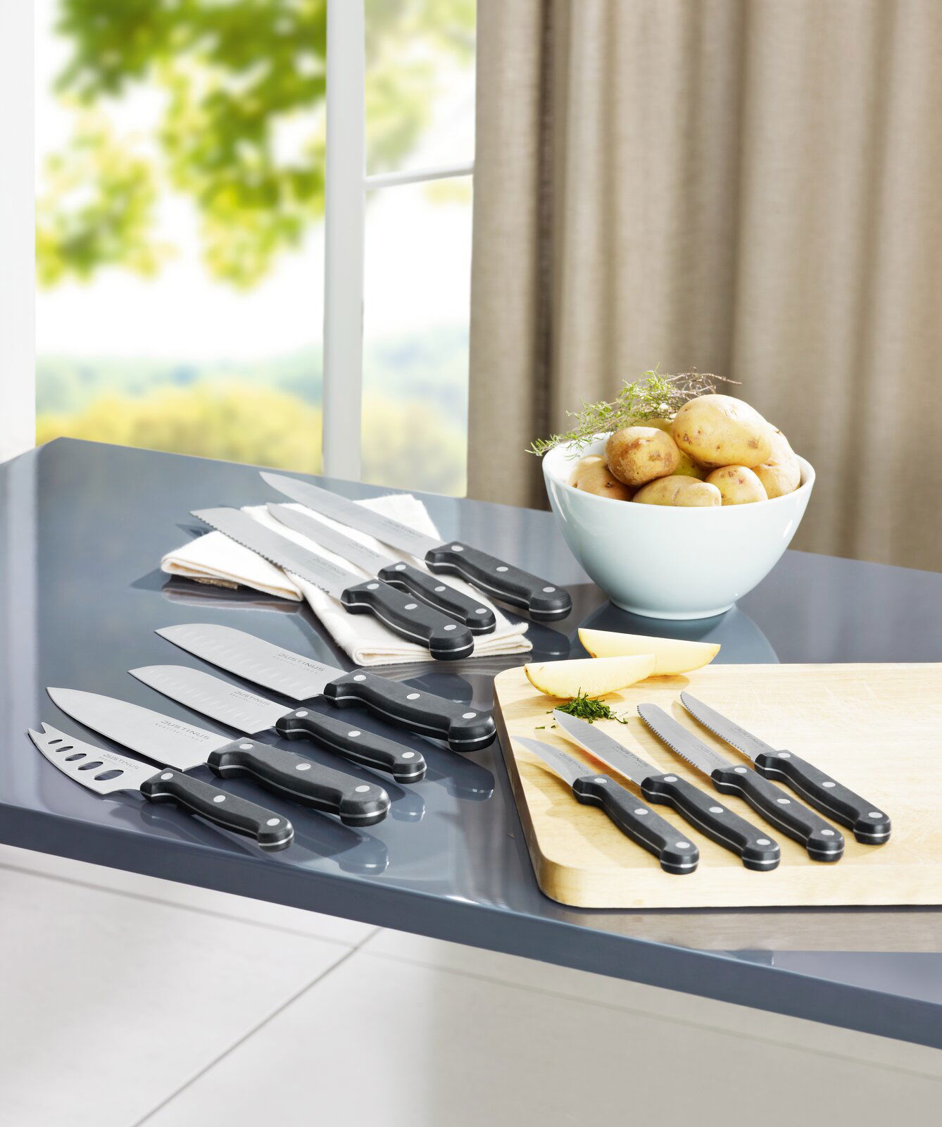 Justinus Brötchenmesser Küchenchef KÜCHENCHEF Verschiedene Küchenmesser, darunter ein Brötchenmesser, auf einem Tisch in einer modernen Küche, mit Blick auf ein Fenster im Hintergrund.