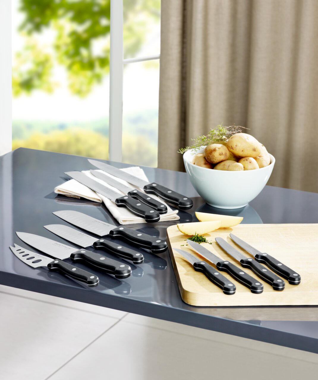 Justinus Brötchenmesser Küchenchef KÜCHENCHEF Verschiedene Küchenmesser, darunter ein Brötchenmesser, auf einem Tisch in einer modernen Küche, mit Blick auf ein Fenster im Hintergrund.