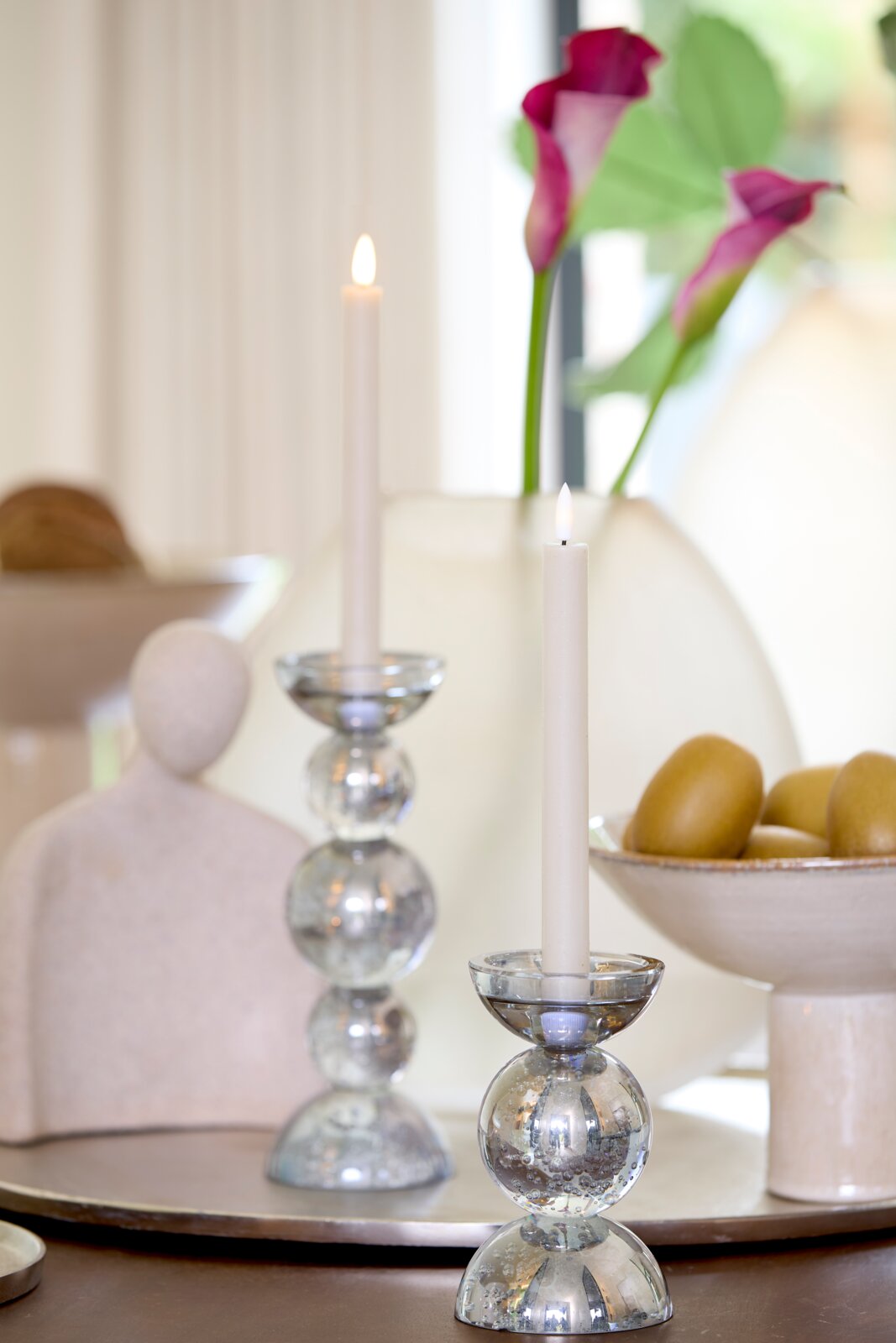Frontaal sfeerbeeld van elegante glazen kandelaars met brandende witte dinerkaarsen, gestyled met roze aronskelken en moderne woonaccessoires.