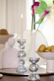 Frontaal sfeerbeeld van elegante glazen kandelaars met brandende witte dinerkaarsen, gestyled met roze aronskelken en moderne woonaccessoires.