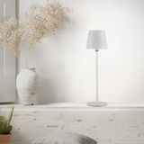 Moderne Akku-Tischleuchte BOTERIA in Weiß, seitliche Perspektive, auf einem rustikalen Regal neben einer Vase mit Trockenblumen.