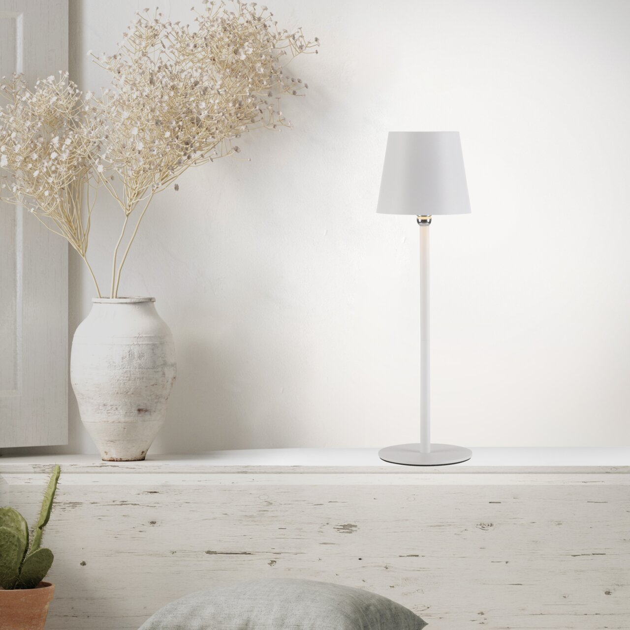 Moderne accu-tafellamp BOTERIA in wit, zijaanzicht, op een rustieke plank naast een vaas met gedroogde bloemen.