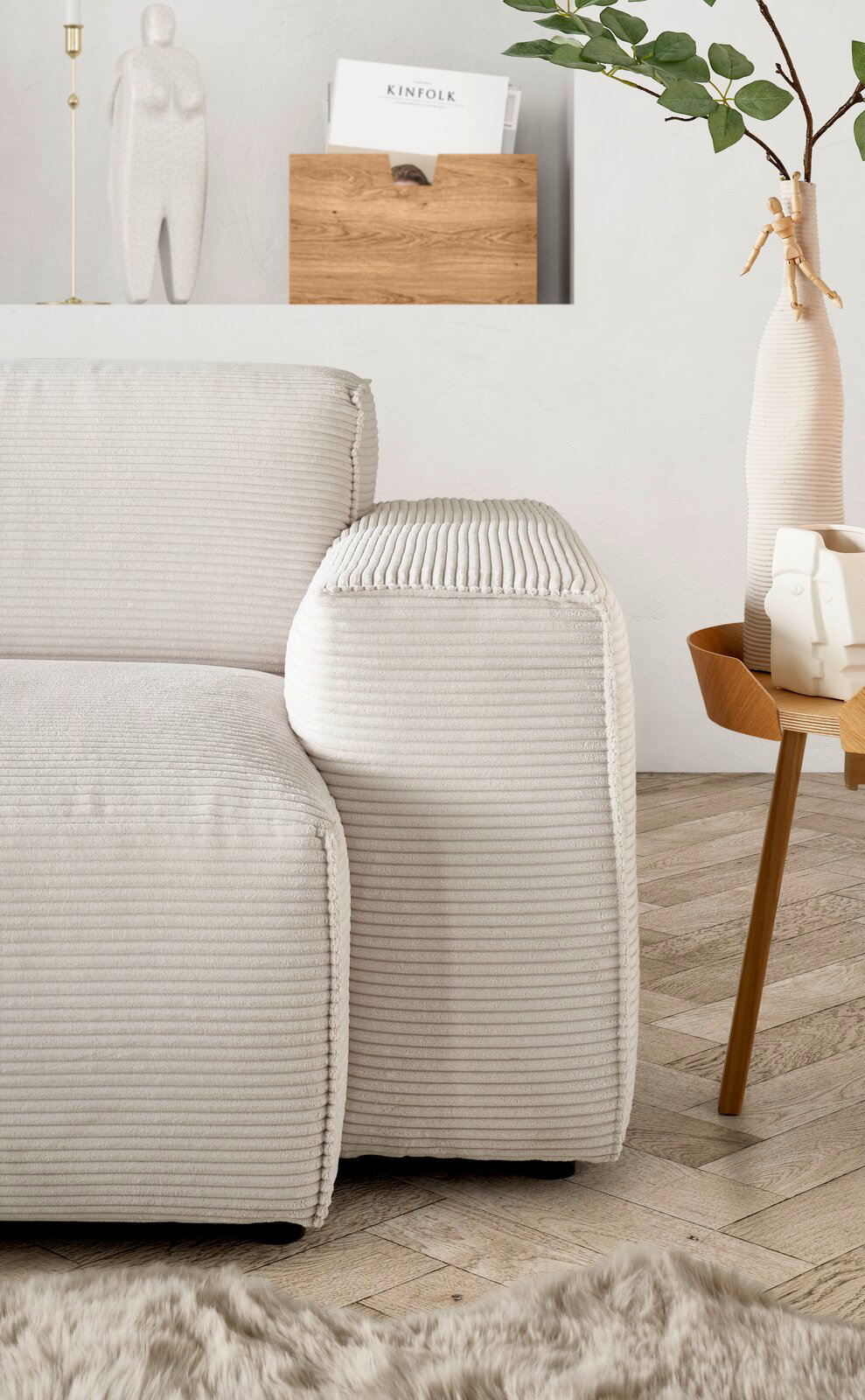 Nahaufnahme einer modernen, hellen Rundecke mit geripptem Stoffbezug, von der Seite betrachtet. Im Hintergrund sind eine Vase mit Zweigen und ein Beistelltisch zu sehen.