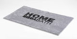 Grauer rechteckiger Badteppich mit der Aufschrift 'HOME BATH' in schwarzer Schrift, perspektivisch von der Seite fotografiert.
