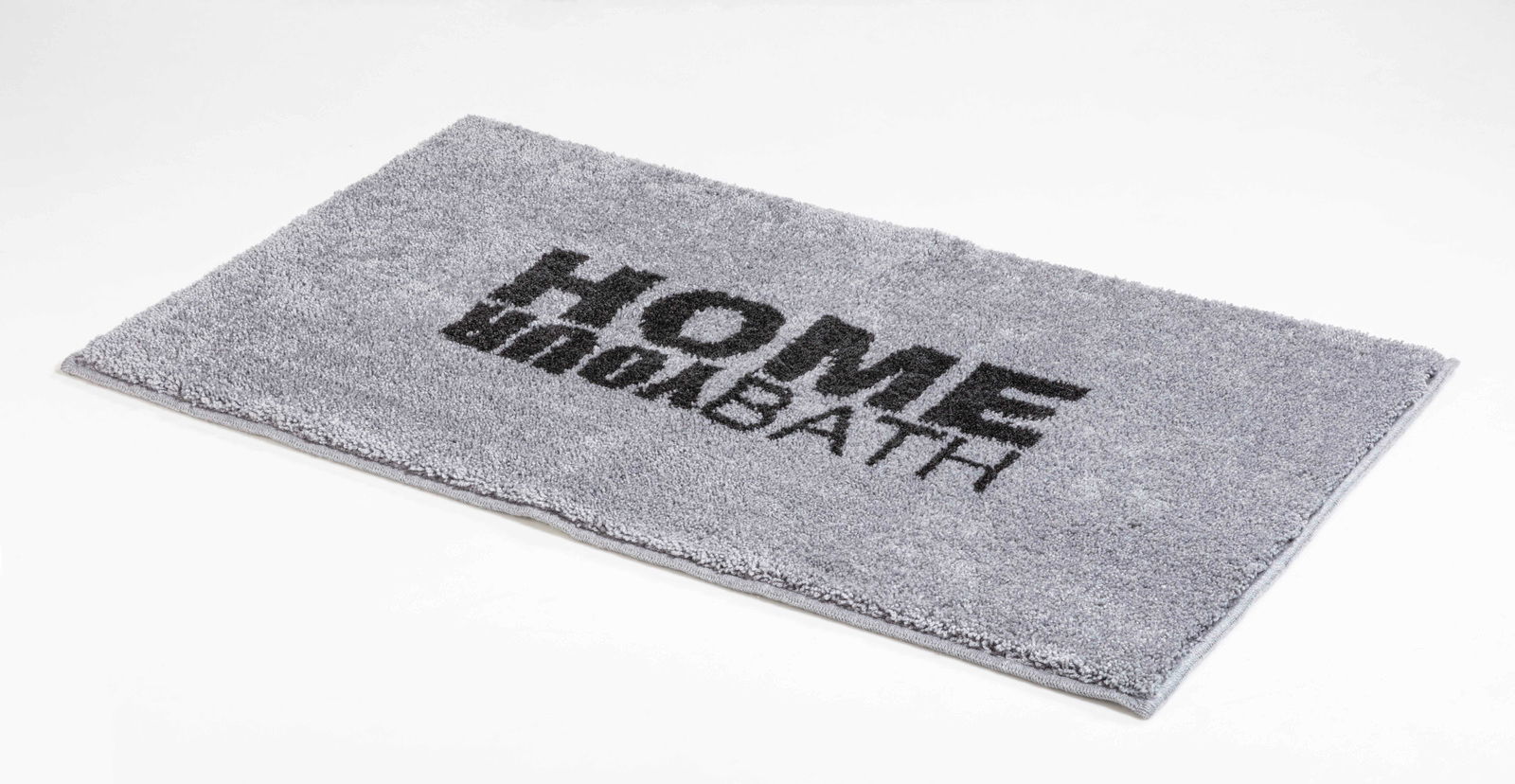Grauer rechteckiger Badteppich mit der Aufschrift 'HOME BATH' in schwarzer Schrift, perspektivisch von der Seite fotografiert.