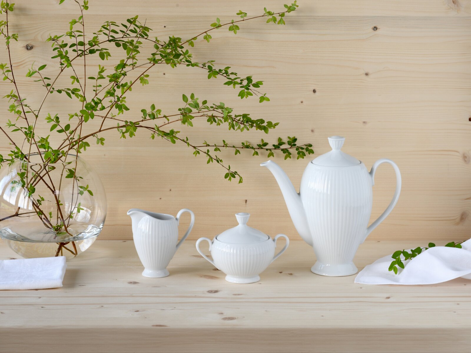 Creatable Kaffeegarnitur 3-tlg VALENCIA Weiße geriffelte Porzellan-Kaffeekanne mit passender Zuckerdose und Milchkännchen auf hellem Holztisch, seitliche Frontansicht auf das Set, dekoriert mit Glasvase und grünen Zweigen.