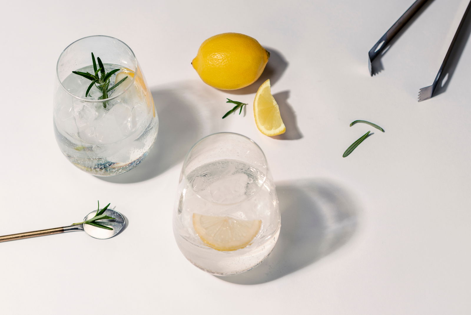 SCHOTT ZWIESEL Gin Tonic Gläser 4-tlg. BAR SPECIAL Zwei Gingläser mit Eis, Zitrone und Rosmarin aus der Vogelperspektive, umgeben von einer ganzen Zitrone, einem Zitronenschnitz, Rosmarinzweigen und einer Zange.