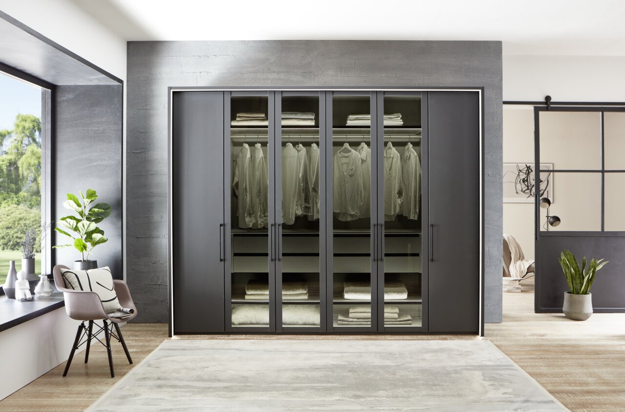 Moderner 6-türiger Drehtürenschrank in dunklem Design, frontal fotografiert, mit Glaseinsätzen und sichtbarer Kleidung und Bettwäsche im Inneren.