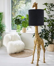 Stehleuchte in Form einer goldenen Giraffe mit schwarzem Lampenschirm, seitliche Perspektive, umgeben von Pflanzen in einem hellen Raum.