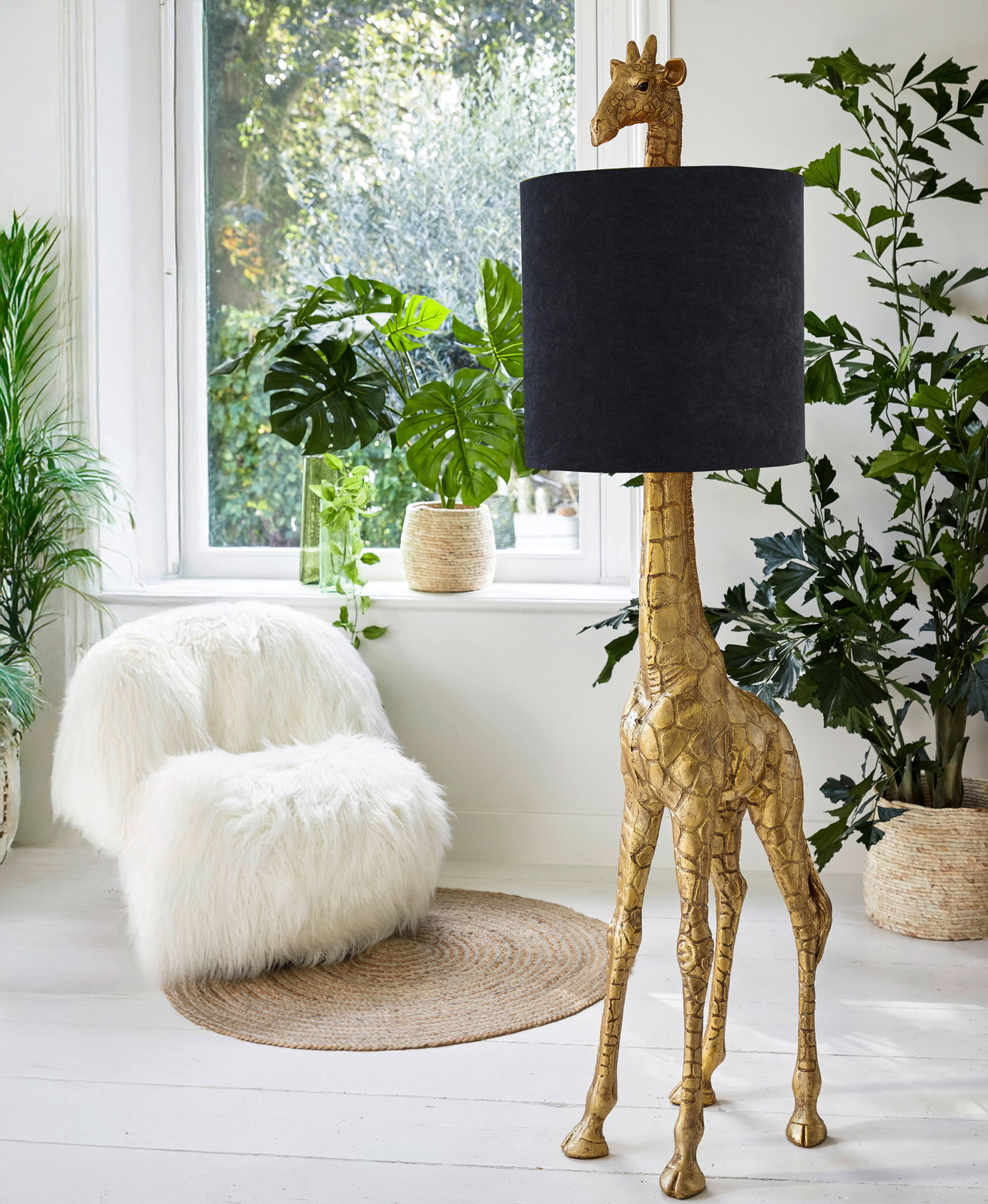Stehleuchte in Form einer goldenen Giraffe mit schwarzem Lampenschirm, seitliche Perspektive, umgeben von Pflanzen in einem hellen Raum.