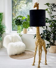 Stehleuchte in Form einer goldenen Giraffe mit schwarzem Lampenschirm, seitliche Perspektive, umgeben von Pflanzen in einem hellen Raum.