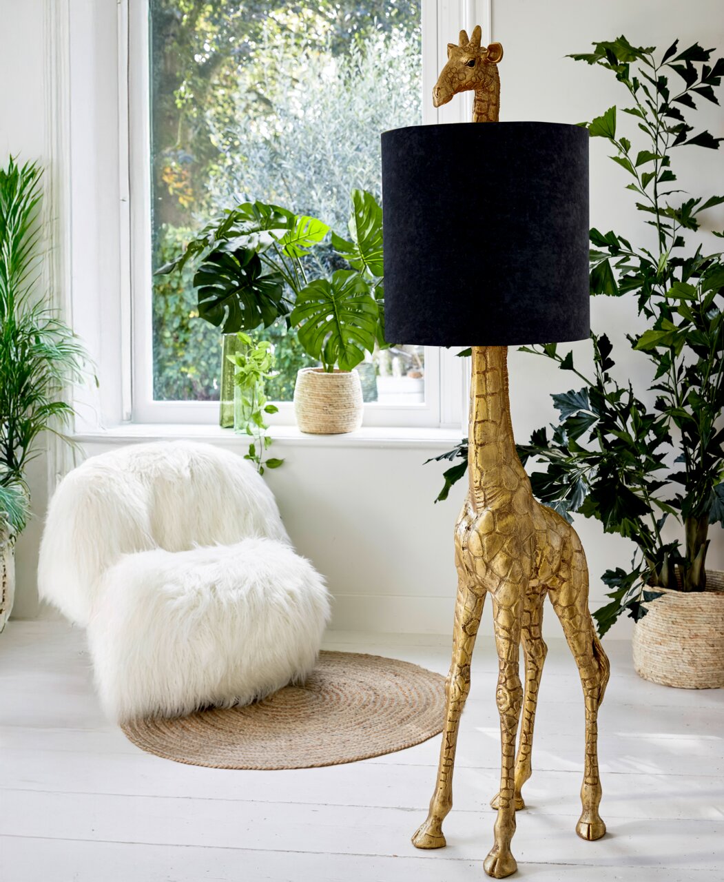 Stehleuchte in Form einer goldenen Giraffe mit schwarzem Lampenschirm, seitliche Perspektive, umgeben von Pflanzen in einem hellen Raum.