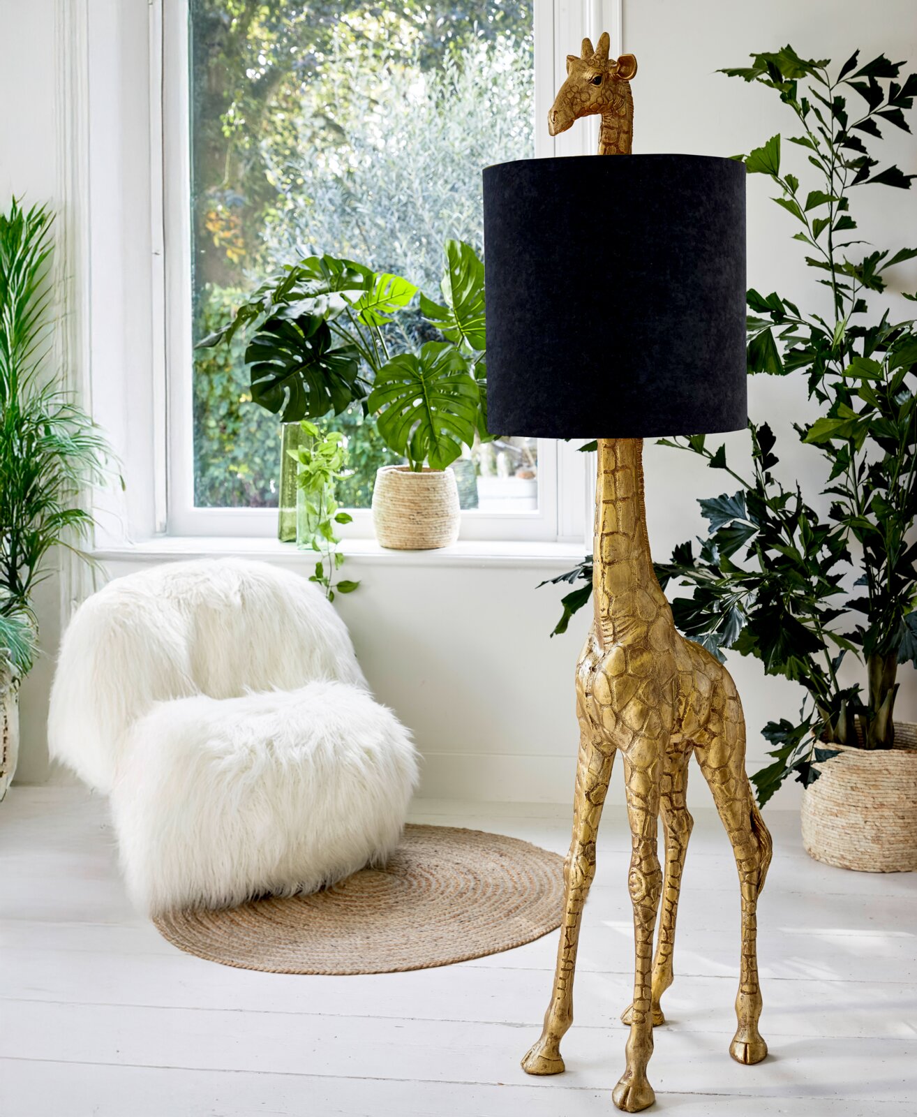Stehleuchte in Form einer goldenen Giraffe mit schwarzem Lampenschirm, seitliche Perspektive, umgeben von Pflanzen in einem hellen Raum.