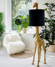 Staande lamp in de vorm van een gouden giraf met een zwarte lampenkap, gezien vanuit de zijkant, omringd door planten in een lichte ruimte.