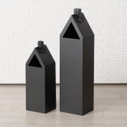 Twee zwarte, huisvormige metalen lantaarns van verschillende grootte met een driehoekige opening en een handvat, vrijstaand op een lichte ondergrond voor een witte bakstenen muur, vooraanzicht licht schuin van voren.